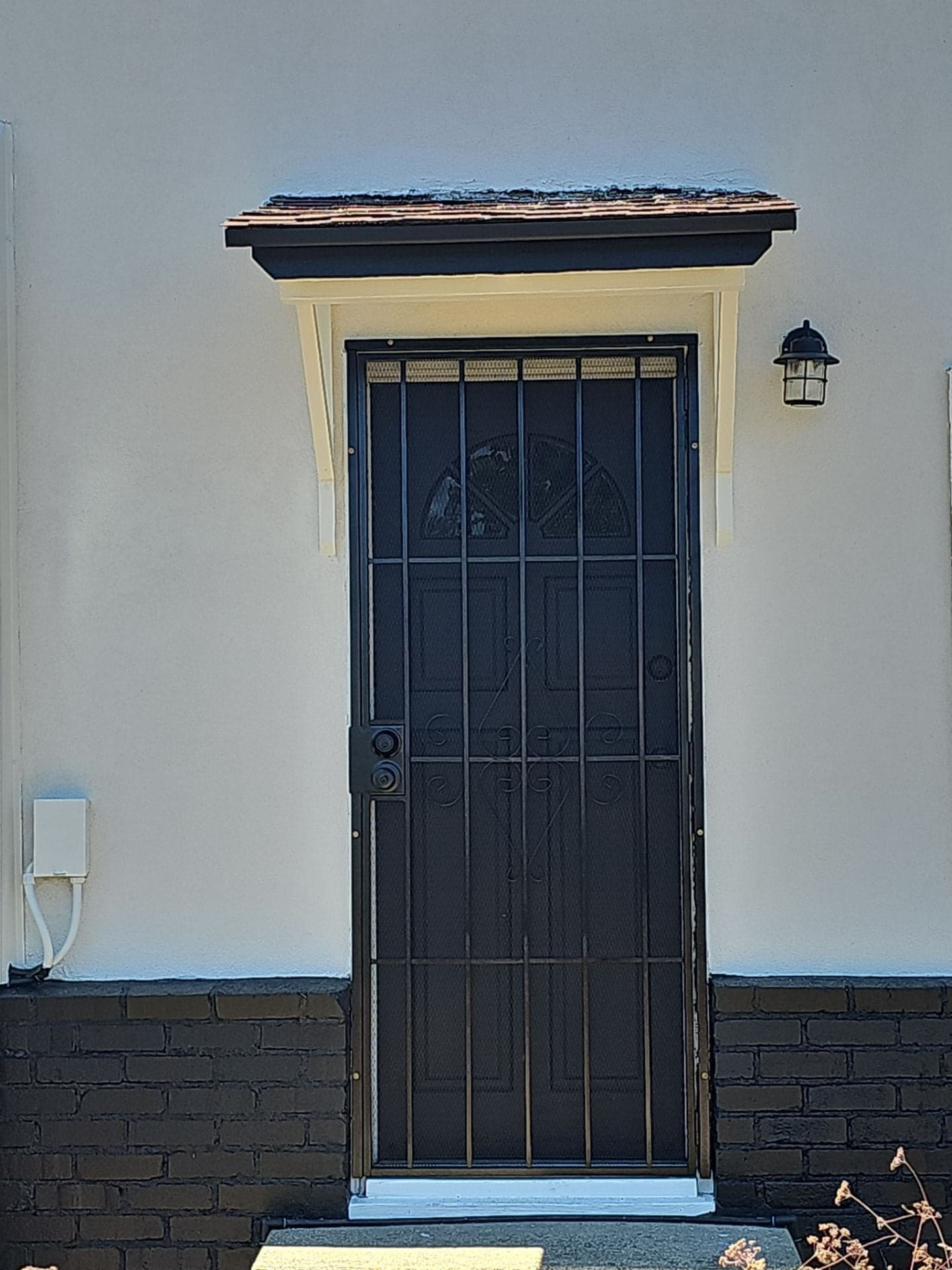 Finished front entry detail by RLS Custom Carpentry showing a paneled door with iron security screen, a custom wood awning, fresh white stucco, and painted brick wainscot on a Mendocino County home