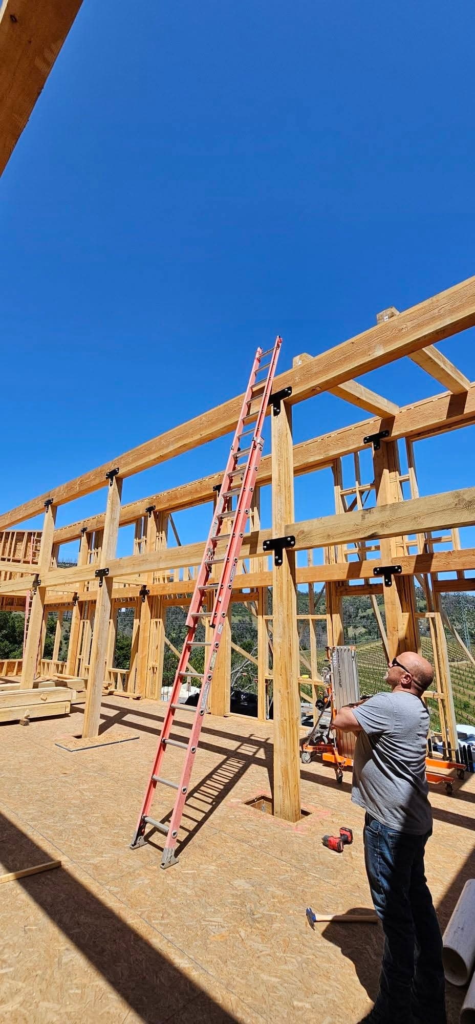 Heavy-timber post-and-beam structural framing of a large building with a craftsman on site, vineyard hills beyond, in Mendocino County California