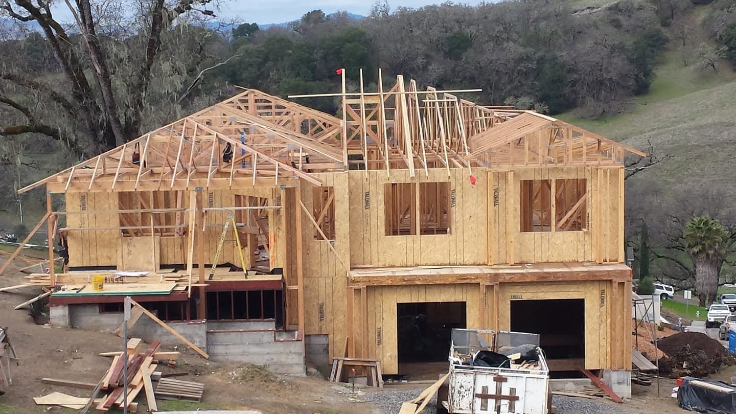 Large two-story custom home under construction with completed roof truss framing and sheathing against a green Mendocino County hillside