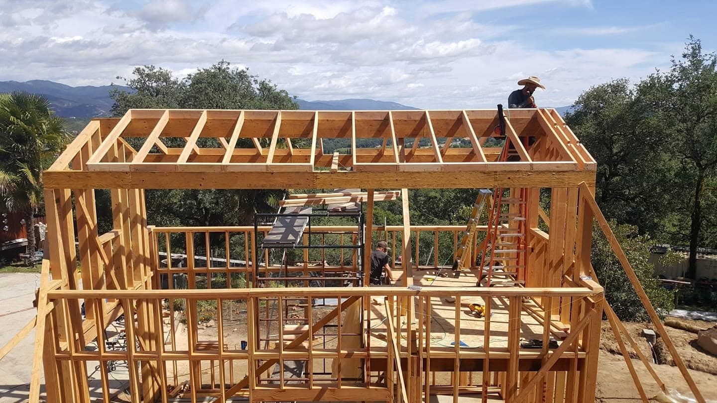 Two-story wood frame home with roof rafters under construction and a framer working on the roofline, mountain views in Mendocino County, CA